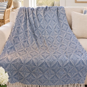 May include: A woven throw blanket featuring a repeating blue and white diamond pattern with intricate geometric details. The blanket has a textured surface and is finished with a fringe trim along the bottom edge, creating a soft and layered appearance.