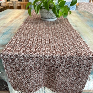 May include: A brown and white knitted table runner with a fringed edge. The runner is placed on a distressed wooden table. A potted plant with green leaves sits on the runner.