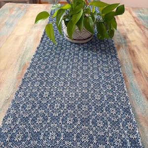 May include: A blue and white patterned table runner with a geometric design. The runner is placed on a distressed wooden table. A potted plant with green leaves sits on top of the runner.