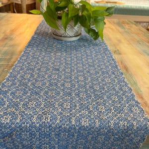 May include: A blue and white patterned table runner with a fringed edge. The runner is made of woven fabric and is placed on a wooden table. A potted plant with green leaves sits on the runner.