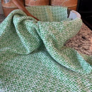 May include: A green and white patterned kitchen towel draped over a white bowl with a wooden spoon inside. The towel has a diamond pattern. Behind the bowl are three ceramic jars with lids.