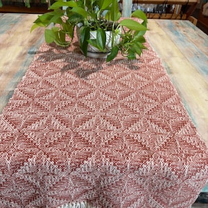 May include: A rectangular woven table runner featuring a repeating diamond pattern in terracotta orange and cream white. The fabric has a textured weave and is finished with a fringe of cream-colored threads along the short edges of the runner.