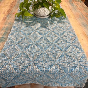 May include: A rectangular table runner featuring a woven geometric diamond pattern in bright turquoise blue and off-white. The textile has a textured appearance with a repeating motif across the surface, providing a consistent color scheme.