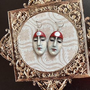 May include: A pair of artisan earrings depicting oval faces with closed eyes and red lips. Each earring has a red cap with a silver circle. The earrings are displayed on a patterned background within a gold-coloured ornate frame.