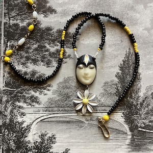 Könnte beinhalten: Eine Halskette mit einem schwarzen und weißen Gesicht als Anhänger und einem gelben Gänseblümchen-Anhänger. Die Halskette besteht aus schwarzen und gelben Perlen.