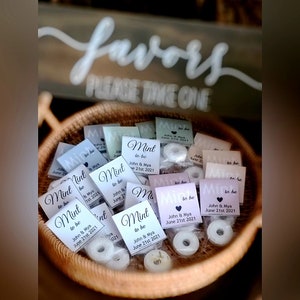 Op de afbeelding: Een mand gevuld met kleine witte pepermuntjes verpakt in doorzichtige cellofaan zakjes met gepersonaliseerde etiketten waarop "Mint to be" staat met de namen van het paar en de datum van de bruiloft.