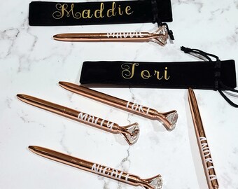 Puede incluir: Bolígrafos de oro rosa con detalles en forma de diamante y nombres personalizados en letras blancas. Cada bolígrafo viene con una bolsa de terciopelo negro, también personalizada con letras doradas. Los nombres "Maddie", "Tori", "Luke", "Macy", "Nicole" y "Andrea" son visibles.