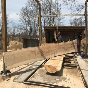 May include: A large wooden slab is being lifted by a crane in a woodworking shop. The slab is light brown in color and has a natural edge. The slab is being lifted over a metal frame and a concrete floor.