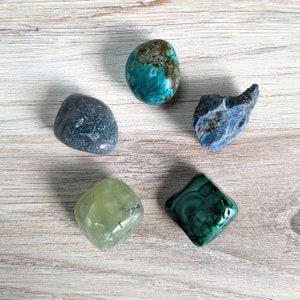 May include: Five tumbled stones in various shades of green and blue. The stones are arranged in a circle on a light wood surface.