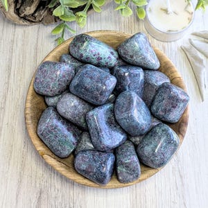 May include: A collection of tumbled green and red ruby zoisite stones in a wooden bowl. The stones are smooth and polished, with a variety of shapes and sizes.