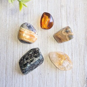 May include: Five polished gemstones of various shapes and colors are arranged on a light wood surface. The stones include amber, banded agate, and a dark green stone with black speckles. The gemstones are likely used for healing or decorative purposes.