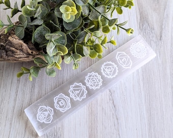 May include: A rectangular, translucent selenite wand with seven white geometric chakra symbols etched along its surface. The wand is displayed on a light wood-grain surface, with green foliage in the background.
