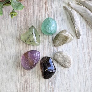 May include: A collection of seven tumbled stones in various colors and textures, including green, purple, brown, and grey. The stones are arranged in a circle on a light wood surface.