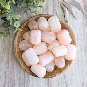 May include: A wooden bowl filled with tumbled peach selenite crystals. The crystals have a smooth, rounded shape and a soft, translucent appearance. The bowl is set on a light-colored wooden surface, with green leaves and dried sage in the background.