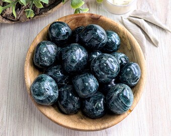 May include: A wooden bowl filled with polished, dark green stones. The stones have a mottled appearance and a smooth, rounded shape. The bowl is made of light-colored wood and sits on a light-colored wooden surface.