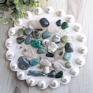 May include: A collection of polished gemstones in various colors, including green, clear, and blue, arranged on a white, scalloped-edge tray. The tray has decorative white and brown accents. The gemstones vary in size and shape, creating a natural, earthy aesthetic.