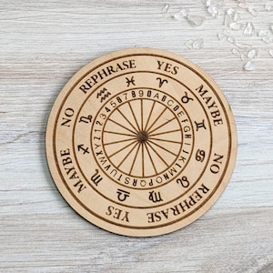 May include: A round wooden planchette with engraved text and symbols. The planchette features a central wheel with numbers and letters, surrounded by phrases like "YES", "NO", and "MAYBE", and zodiac symbols. The planchette is made of light-colored wood.