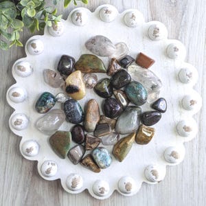 May include: A collection of polished gemstones in various colors, including green, brown, clear, and black, arranged on a white, scalloped tray with decorative white accents. The gemstones vary in size and shape, creating a natural, earthy aesthetic.