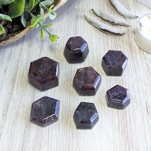 May include: Seven dark red, hexagonal, polished, gemstone points on a light wood surface.