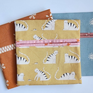 May include: Three fabric pouches in shades of orange, yellow, and blue. The yellow pouch features a repeating pattern of white tigers with black stripes. The orange pouch has a floral design, and the blue pouch has a sun pattern.