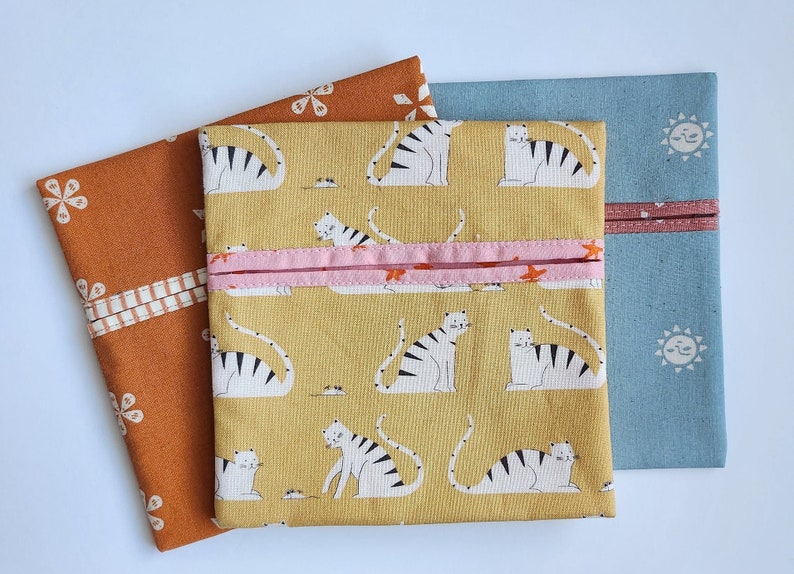 May include: Three fabric pouches in shades of orange, yellow, and blue. The yellow pouch features a repeating pattern of white tigers with black stripes. The orange pouch has a floral design, and the blue pouch has a sun pattern.
