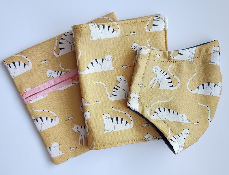 A set of matching mask, mask pouch, and vaccine cardholder. The fabric on each item is yellow with white cats playing on it.