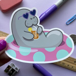 May include: A gray manatee wearing heart-shaped sunglasses and holding a glass of iced tea, relaxing in a pink and blue polka dot inflatable pool float.