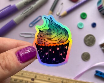 May include: A holographic sticker of a rainbow cupcake with a black cupcake liner and white stars. The frosting is a swirl of rainbow colors with a green swirl on top.