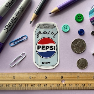 A sticker of a cartoon of a light blue can of Diet Pepsi with the text "fueled by" in the top corner is shown on a purple background surrounded by small objects.