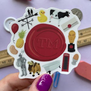 May include: A colorful sticker with a red seal design and the letters "TM" in the center. The sticker features various illustrations, including a pineapple, a cow, a camel, and a red telephone booth.