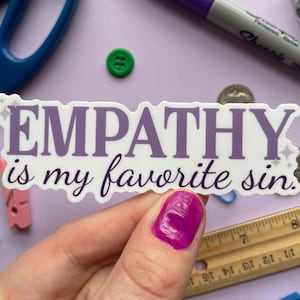 A sticker of purple text that reads "empathy is my favorite sin" is held above a purple background covered in small objects.