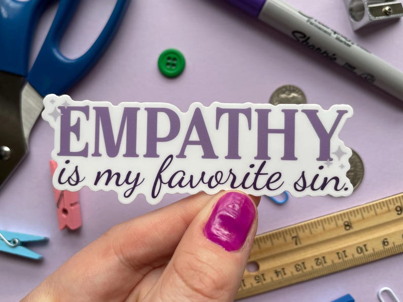 A sticker of purple text that reads "empathy is my favorite sin" is held above a purple background covered in small objects.