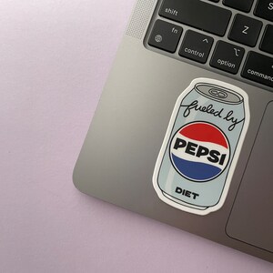 A sticker of a cartoon of a light blue can of Diet Pepsi with the text "fueled by" in the top corner is shown on a silver laptop.