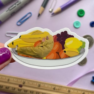 May include: A white ceramic bowl filled with a variety of fruit, including bananas, grapes, and oranges. A person is laying inside the bowl, surrounded by the fruit.