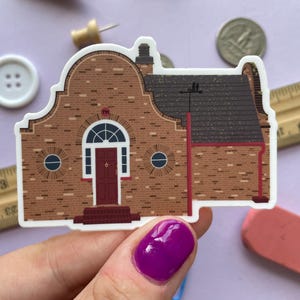 May include: A sticker featuring a detailed illustration of a brick building with a dark red door, arched window, and a dark gray roof. The building has two round windows and a chimney. The sticker is held by a hand with purple nail polish.