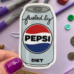 A sticker of a cartoon of a light blue can of Diet Pepsi with the text "fueled by" in the top corner is held over a purple background covered in objects.