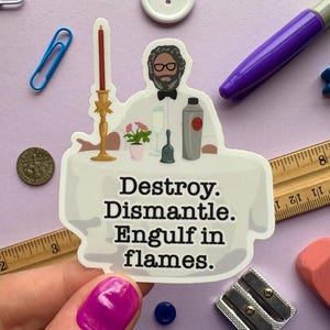 May include: A sticker featuring a cartoon of a man behind a table with the text "Destroy. Dismantle. Engulf in flames." The sticker is surrounded by office supplies, including a ruler, coins, and a purple marker.