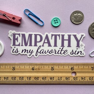 A sticker of purple text that reads "empathy is my favorite sin" is shown on a purple background covered in small objects.