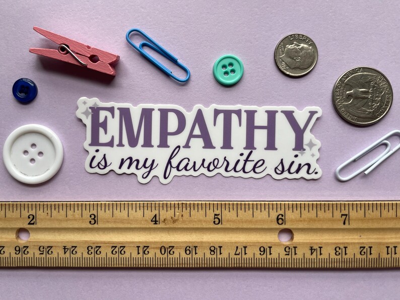 A sticker of purple text that reads "empathy is my favorite sin" is shown on a purple background covered in small objects.