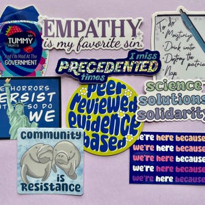 May include: A collection of colorful stickers with various messages and designs. The stickers include phrases like "Empathy is my favorite sin", "The horrors persist but so do we", "Science solutions solidarity", and "Community is resistance".
