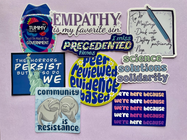 May include: A collection of colorful stickers with various messages and designs. The stickers include phrases like "Empathy is my favorite sin", "The horrors persist but so do we", "Science solutions solidarity", and "Community is resistance".