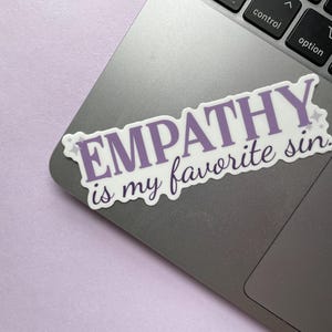 A sticker of purple text that reads "empathy is my favorite sin" is shown on a silver laptop on a purple background.