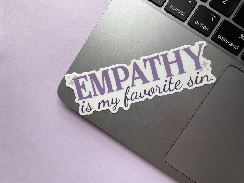 A sticker of purple text that reads "empathy is my favorite sin" is shown on a silver laptop on a purple background.