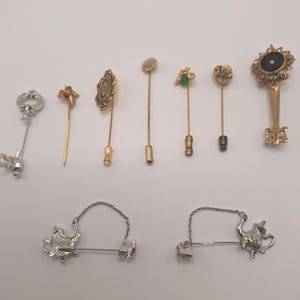 May include: A collection of nine stick pins and lapel pins in various metal finishes including gold and silver tones. The set features diverse designs such as key shapes, floral motifs with small stones, a black circular emblem, and teapot shapes.