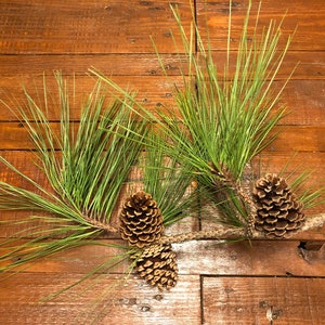 May include: A sprig of pine needles and three pine cones rest on a rustic wooden surface. The long, thin green needles contrast with the brown, textured cones. The wood grain adds a natural, earthy feel.