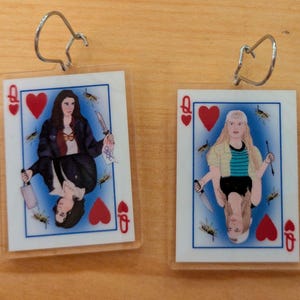 May include: A pair of clear acrylic earrings featuring illustrations of the Queen of Hearts playing card. Each card depicts a woman holding a knife, with a blue background and red heart symbols.