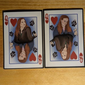 May include: Two playing cards featuring a woman with long brown hair. The cards are the Queen of Hearts. The woman is wearing a brown jacket and a blue shirt. The background is blue with paw prints.