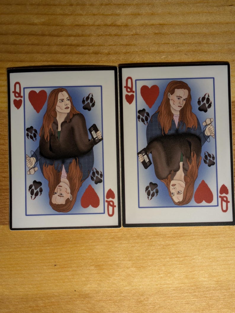 May include: Two playing cards featuring a woman with long brown hair. The cards are the Queen of Hearts. The woman is wearing a brown jacket and a blue shirt. The background is blue with paw prints.