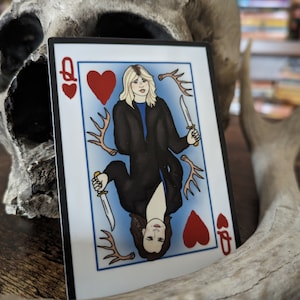 May include: Playing card featuring the Queen of Hearts with two women holding knives. The card has a red heart and antlers in the background.