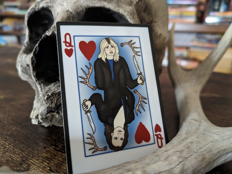 May include: Playing card featuring the Queen of Hearts with two women holding knives. The card has a red heart and antlers in the background.